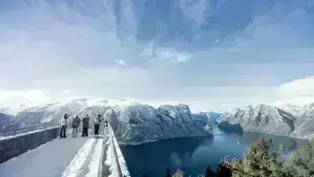 A group stare over a view point in Myrkdalen, looking at a vast landscape of snowy mountains, lush fields and wide, flowing rivers all under a bright blue sky.