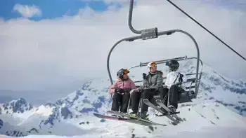 Three skiers sitting side by side on a ski lift, dressed in winter gear, with snow-covered mountains in the background.
