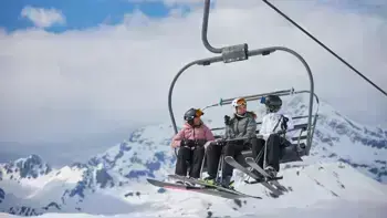 Three skiers sitting side by side on a ski lift, dressed in winter gear, with snow-covered mountains in the background.
